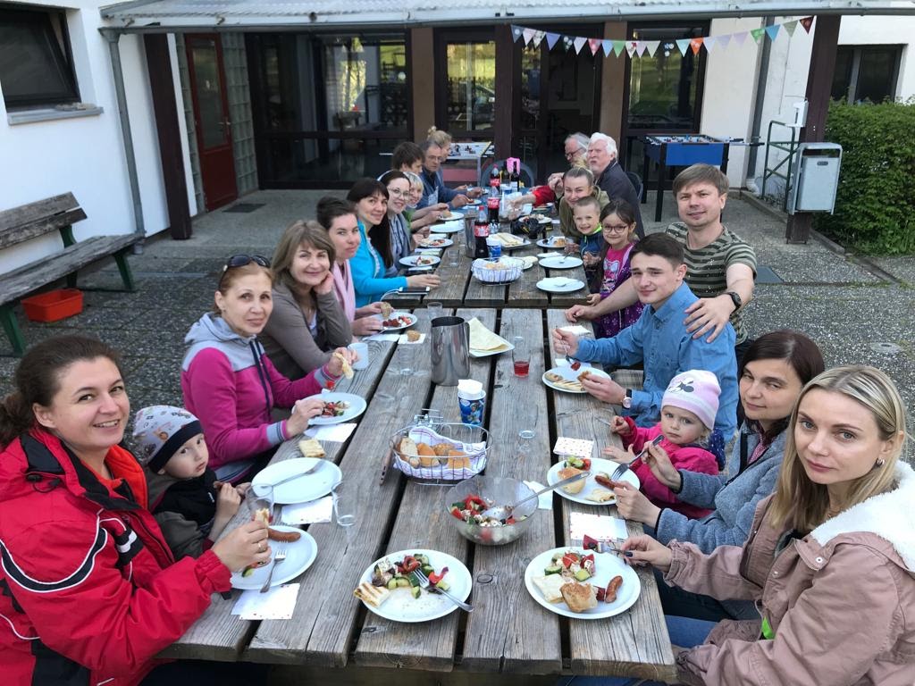 Gemeinsames Essen im Freien: Erwachsene und Kinder sitzen an einem langen Holztisch
